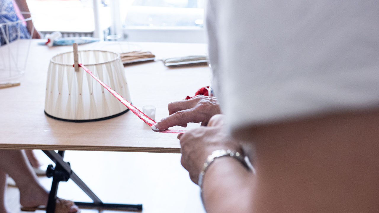 Mesure d’un galon face à un abat-jour plissé posé sur la table, illustrant la préparation minutieuse qui rythme le quotidien d’un abat-jouriste.