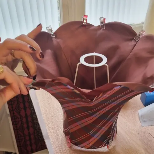 Montage d’un abat-jour aux courbes sculptées, habillé d’un tissu brun et de motifs écossais, en cours d’ajustement par un stagiaire