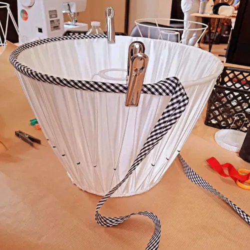 Abat-jour plissé en cours de montage, révélé à travers ses plis réguliers et son ruban posé pour la finition, illustrant l’apprentissage d’un savoir-faire minutieux.