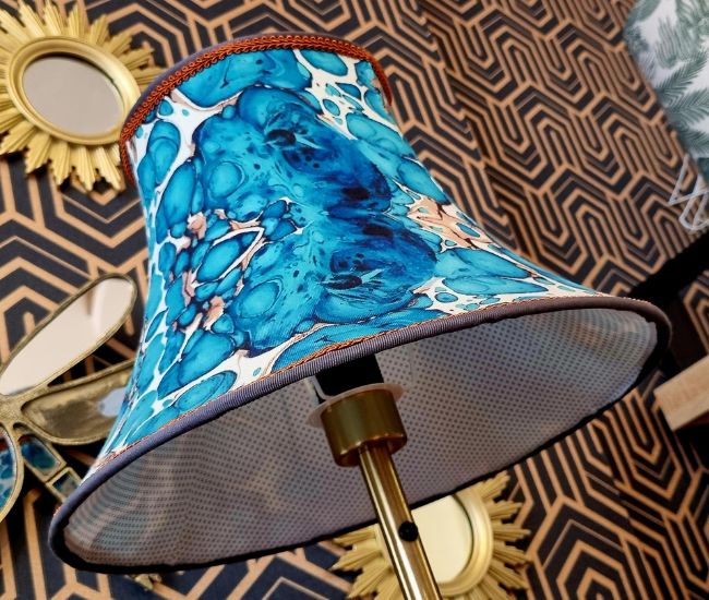 Abat-jour aux motifs marbrés bleus posé sur un pied doré, symbole de la créativité et de la maîtrise artisanale du métier d’abat-jouriste.