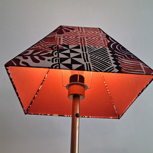 “Abat-jour habillé d’un tissu graphique bicolore, éclairé de l’intérieur par une lumière orange chaleureuse.