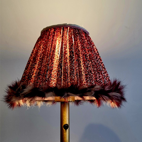 Abat-jour plissé à motifs sombres, bordé d’une frange de plumes, diffusant une lumière chaude.