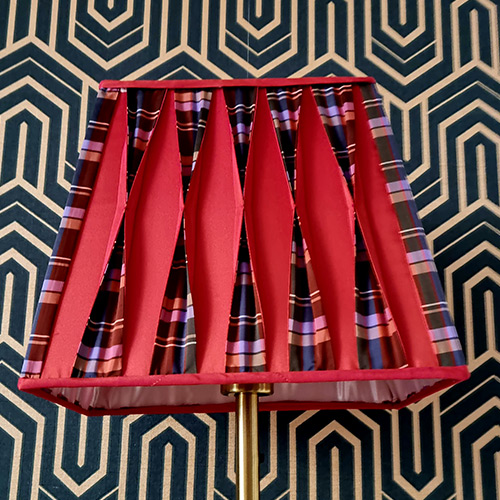 Abat-jour carré mêlant plis rouges et bandes de tissu écossais, créant un motif rythmé et coloré.